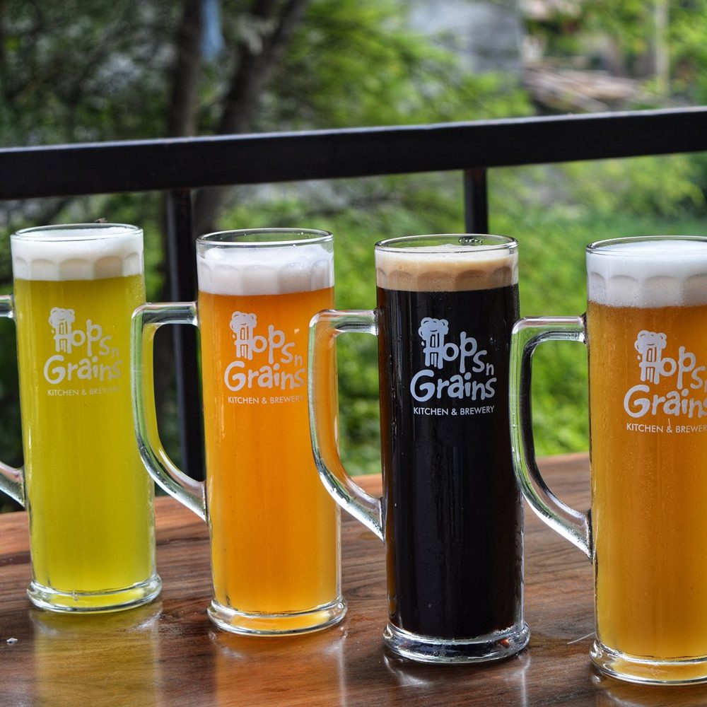 Four Hops n Grains beers, from pale gold to near black, in branded tall mugs on a terrace table